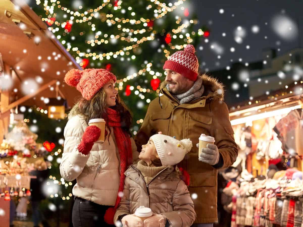 A mom, dad, and daughter each hold a cup of hot cocoa at a Christmas market with holiday lights in the background