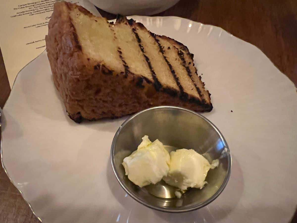 A white plate contains a grilled bannock wedge and a silver cup of butter