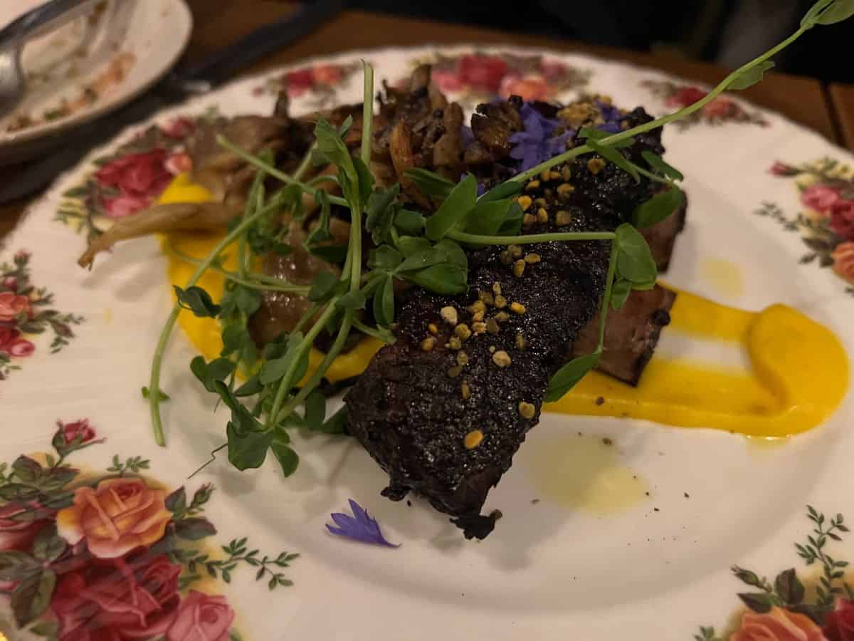 Bison ribs and squash puree topped with greens and purple flower petals on a plate