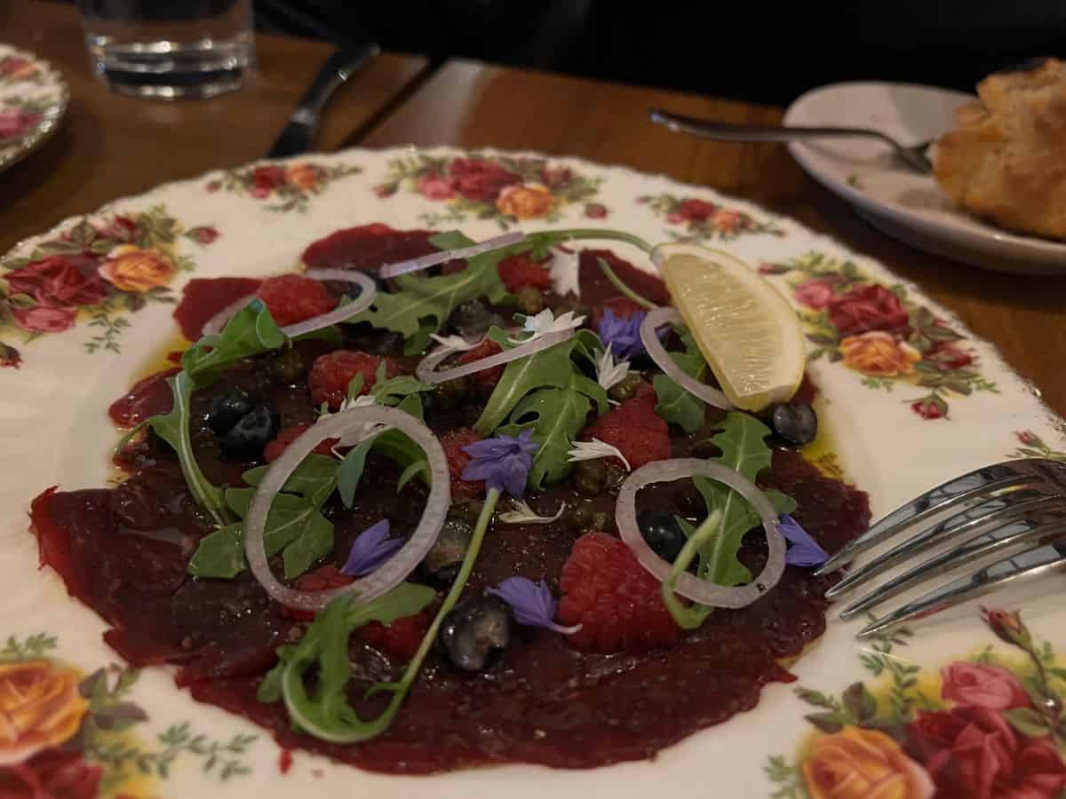 A plate of raw elk topped with berries, greens, and flower petals