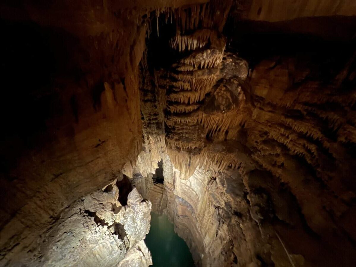 Stalactites hang from a cave ceiling above a lake with turquoise water