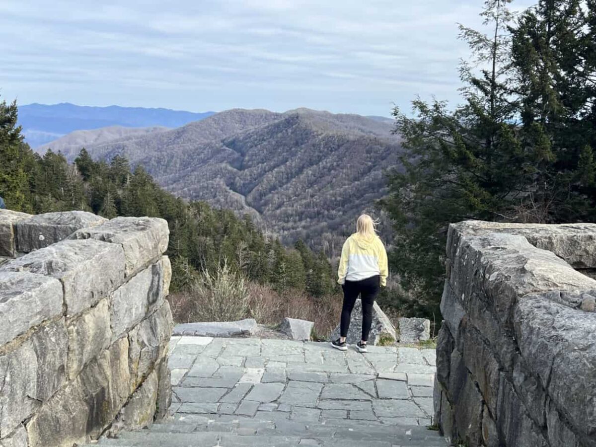 A woman stands at the edge of an overlook to view mountains.