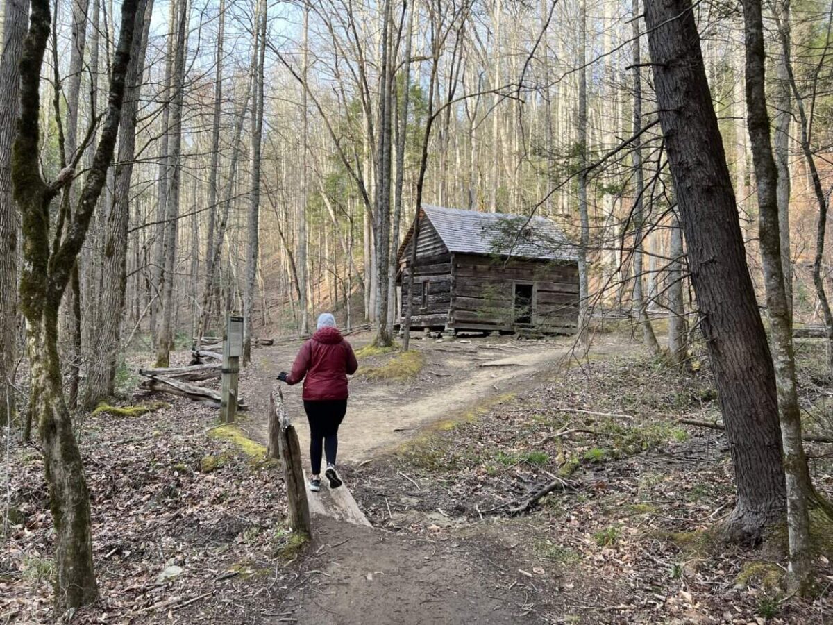 A woman in a maroon coat hikes toward a historic cabin in Great Smoky Mountains National Park.