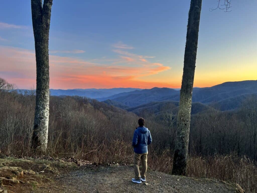 A man stands between two trees and looks ahead at mountains during sunset