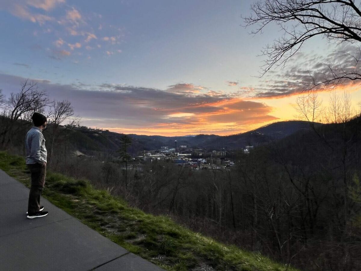 A man enjoys sunrise with views of Gatlinburg below and mountains on the horizon.