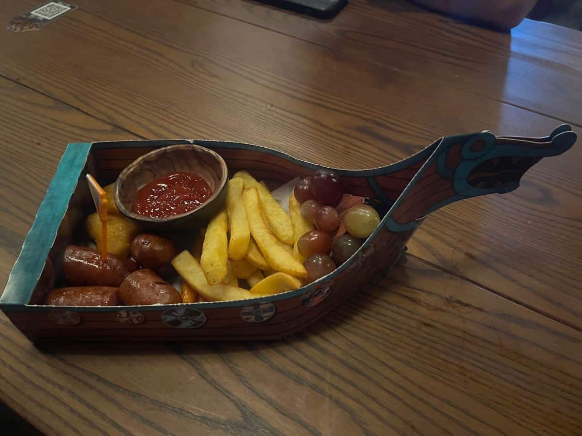 A paper Viking ship holds a meal of mac & cheese bites, sausages, fries, and grapes.