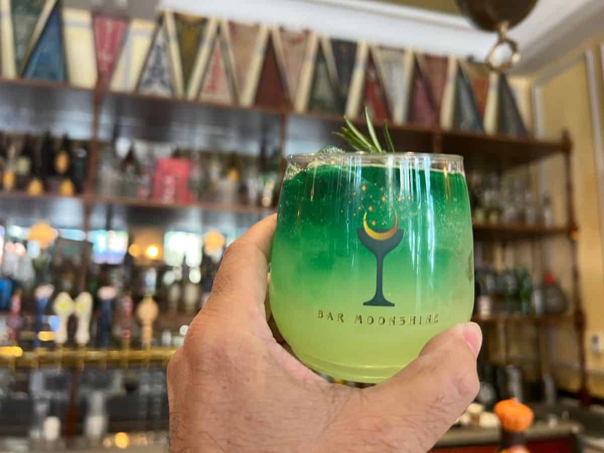A hand holds a glass with a colorful drink with bar memorabilia faded in the background.