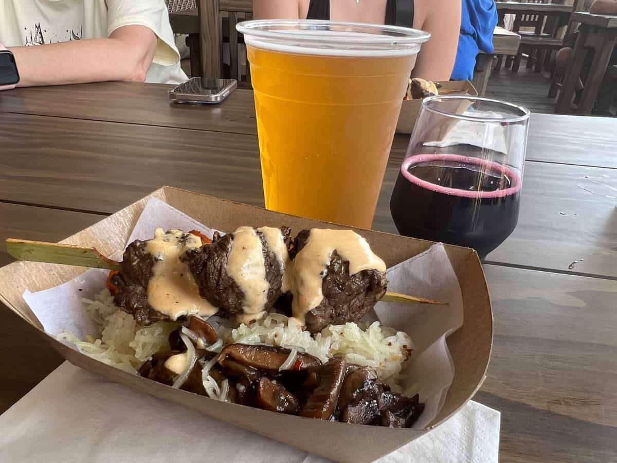 A cardboard carton sits on a table containing a skewer of steak topped with sauce on a bed of rice. A glass of amber beer and a cup of purple mead sit behind the food carton.