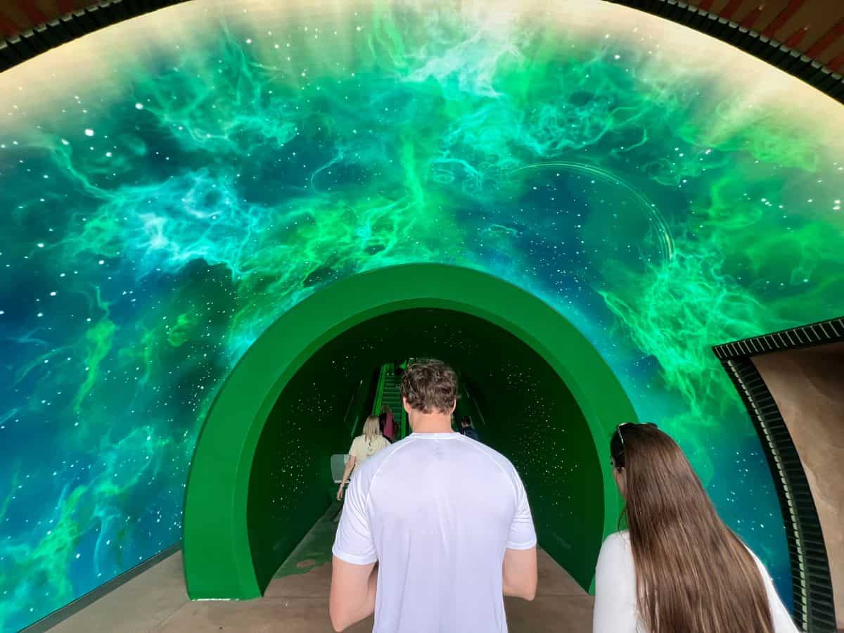 A man and a woman prepare to go up an escalator surrounded by a green arch that resembles a pipe.