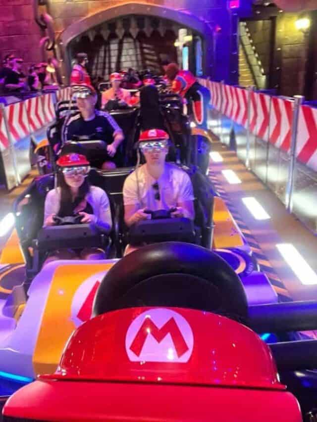Riders sit in their car wearing a Mario hat and goggles on the Mario Kart: Bowser's Challenge ride in Epic Universe.