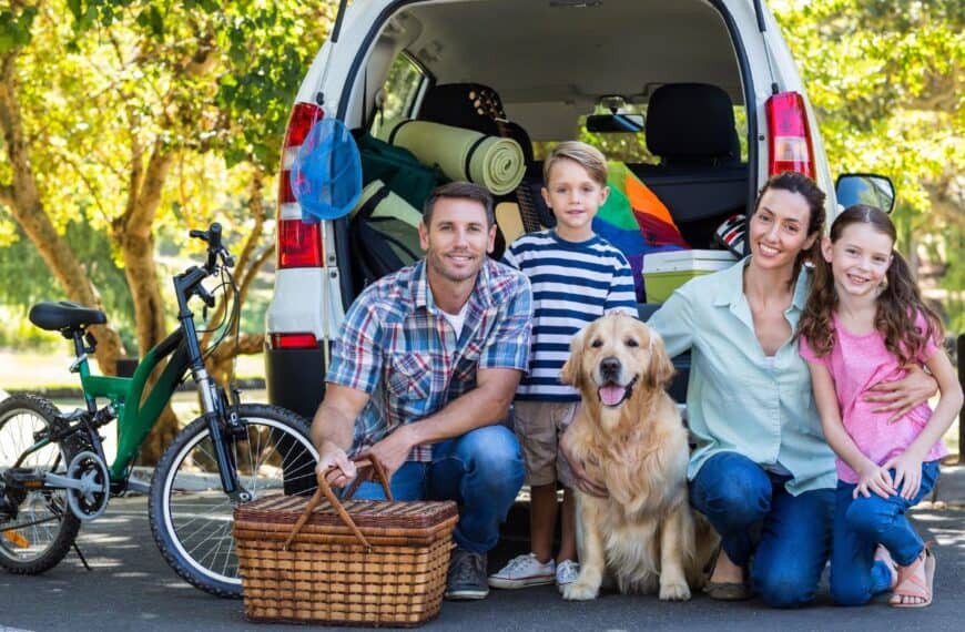 A husband and wife, their young son and young daughter, and family dog kneel behind their vehicle loaded with vacation gear.