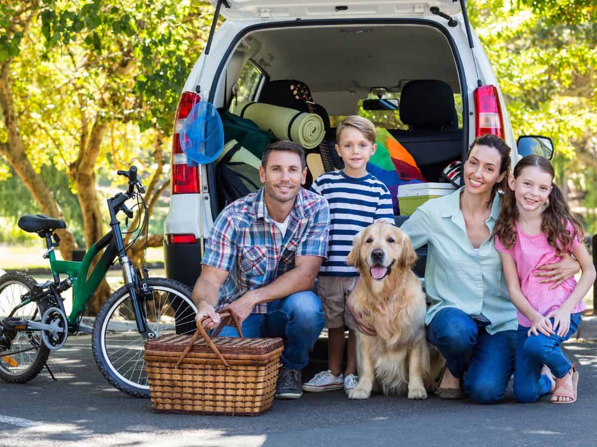 A husband and wife, their young son and young daughter, and family dog kneel behind their vehicle loaded with vacation gear.