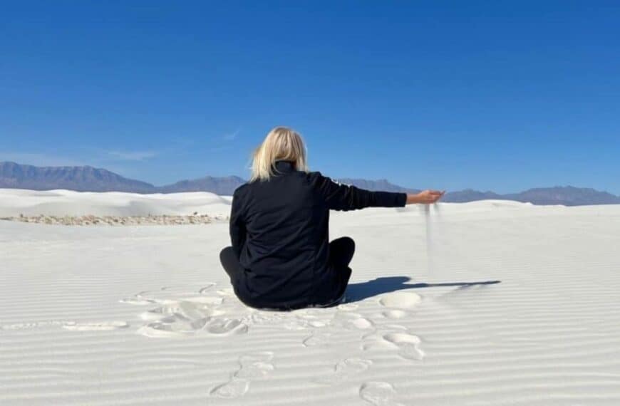 A woman sits in the sand, slowly letting sand sift through her outstretched fingers in White Sands National Park.