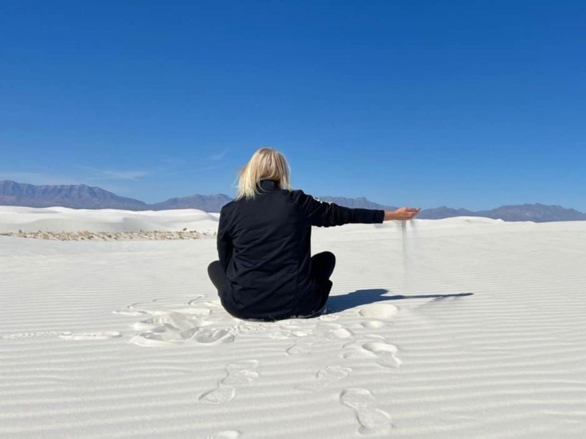 A woman sits in the sand, slowly letting sand sift through her outstretched fingers in White Sands National Park.