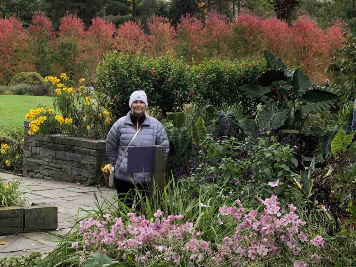 A woman stands next to colorful flowers. Reasons to visit the Finger Lakes, charming small towns like Ithaca, New York.