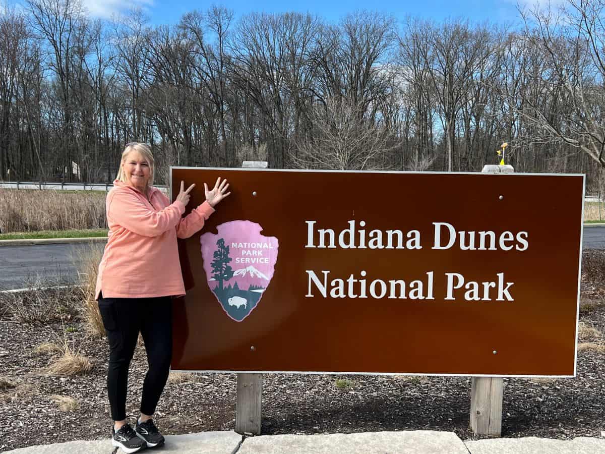 A woman holds up two fingers on one hand and 5 on the other next to the Indiana Dunes National Park sign.