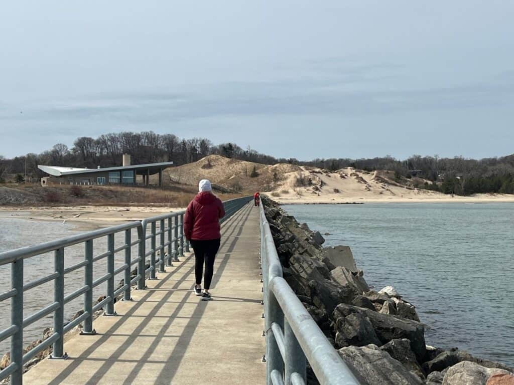 Portage Lakefront is one of the best birding spots in Indiana Dunes National Park. Here, a woman walks along the pier with a pavilion and a sand dune in the distance.