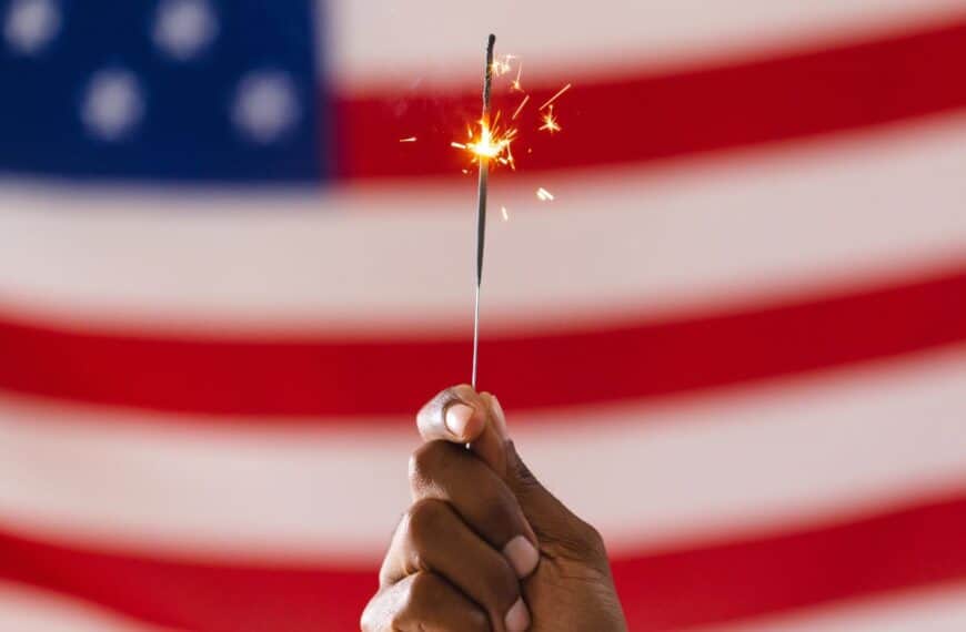 Fizzling sparkler held in front of a faded American flag, symbolizing underwhelming Fourth of July celebrations.