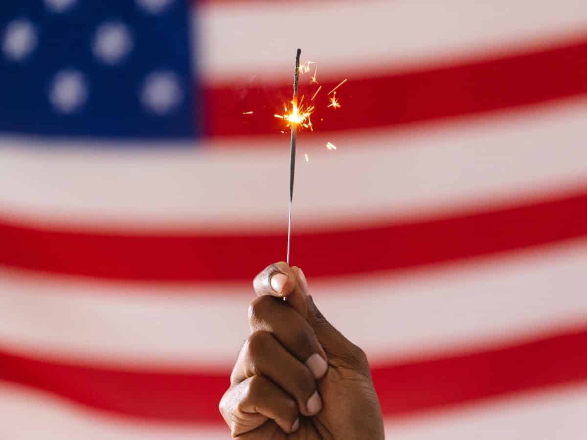 Fizzling sparkler held in front of a faded American flag, symbolizing underwhelming Fourth of July celebrations.