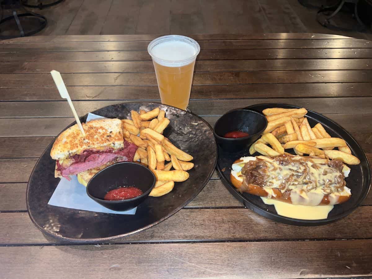 Plates with sandwiches and fries and a cup of beer sit on a wooden table.