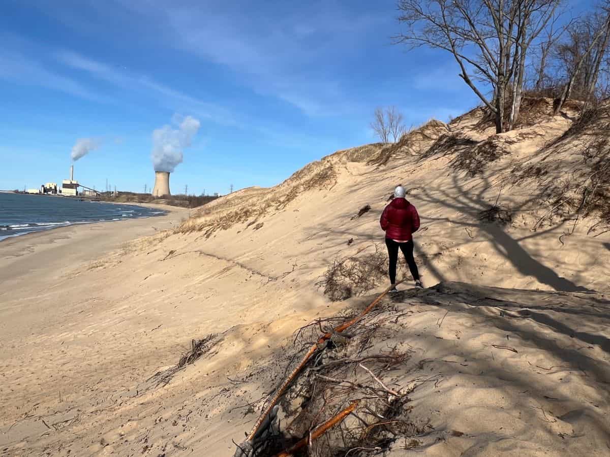 A woman looks at a smokestack from a beach, a sight that makes some guests question if Indiana Dunes National Park is worth visiting.