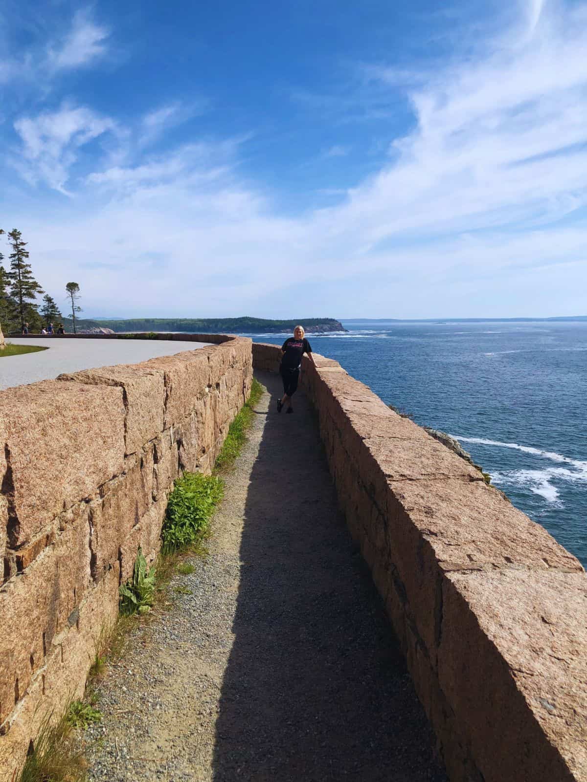 A woman stands along a path between granite walls with a view of the Atlantic Ocean, Acadia National Park, ME