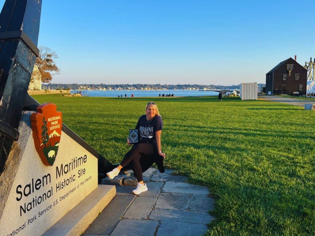 A woman sits next to the Salem Maritime National Historic Site sign in Salem, MA