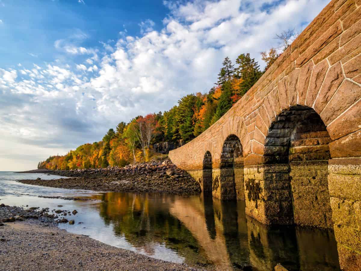 Still waters in Acadia National Park show the reflection of a stone bridge with arches as trees don fall foliage in the background