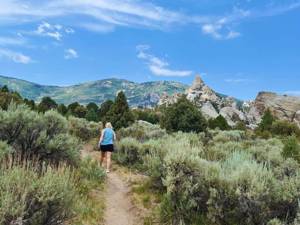 A woman hikes a trail with rocky spires to the right and towering green mountains in the background