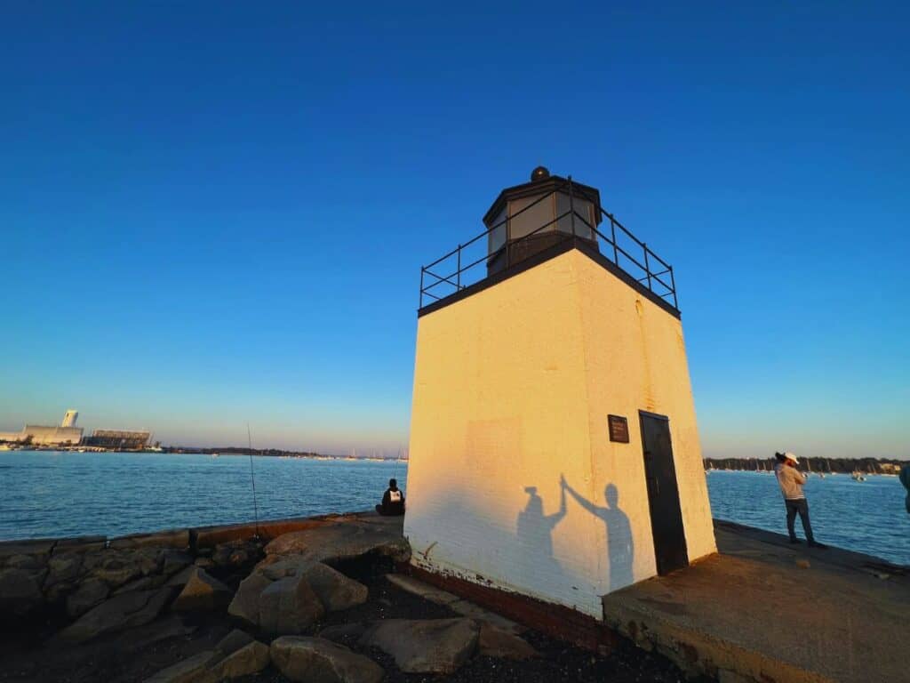 A man and woman give each other a shadow high five against the Derby Wharf Light in Salem, MA