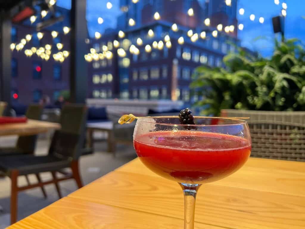 A glass with a red cocktail sits on a table under string lights on a rooftop in downtown Boston
