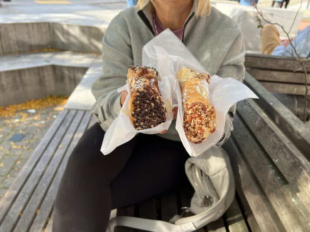 A woman holds two cannoli, one in each hand while sitting on a bench in downtown Boston