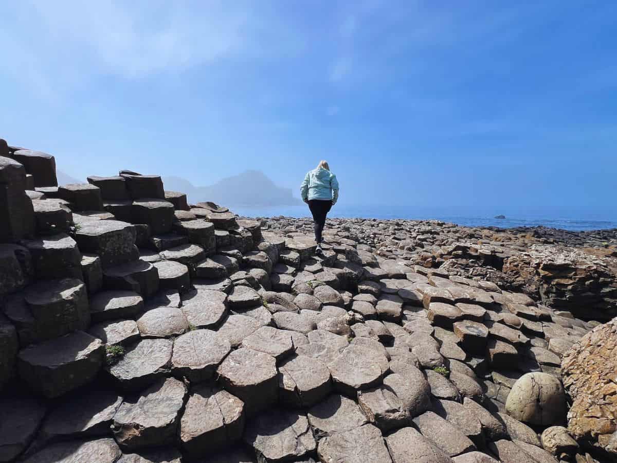 A woman walks across basalt columns at Giant's Causeway with a mountain shrouded in mist in the distance