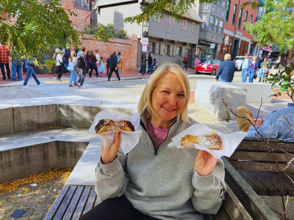 A woman smiles while holding up two cannoli just outside the Paul Revere House on the Freedom Trail