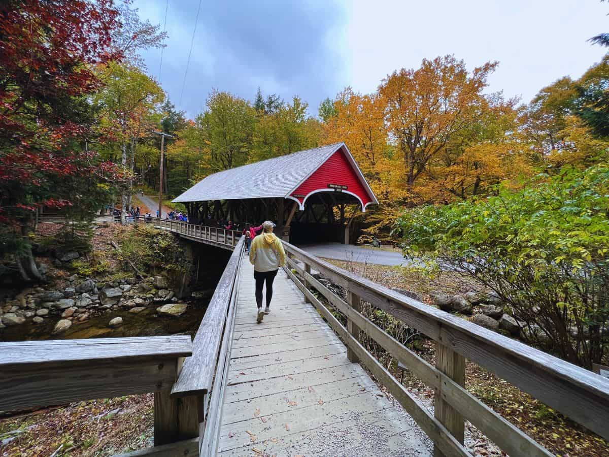 A woman walks a boardwalk trail toward a red covered bridge on the Flume Gorge Trail, NH