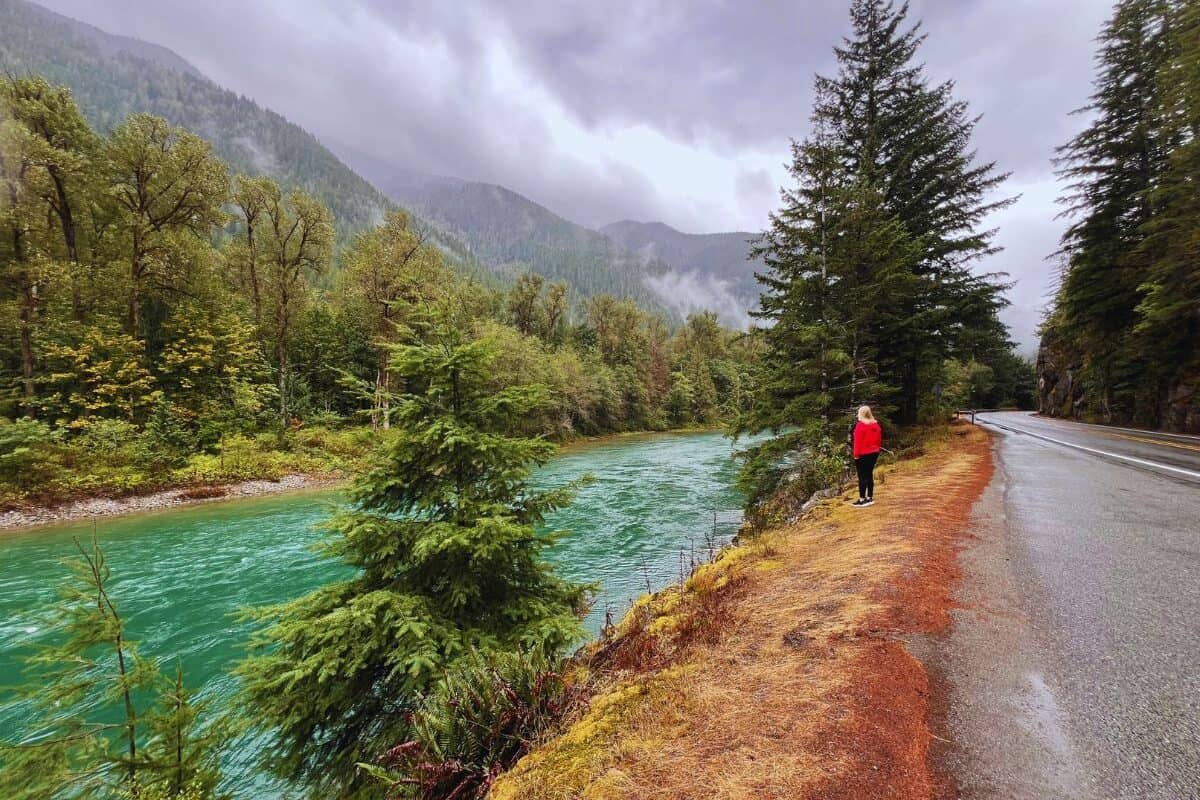 A woman stands just off the edge of the road, looking at the blue waters of the Skagit River in Washington