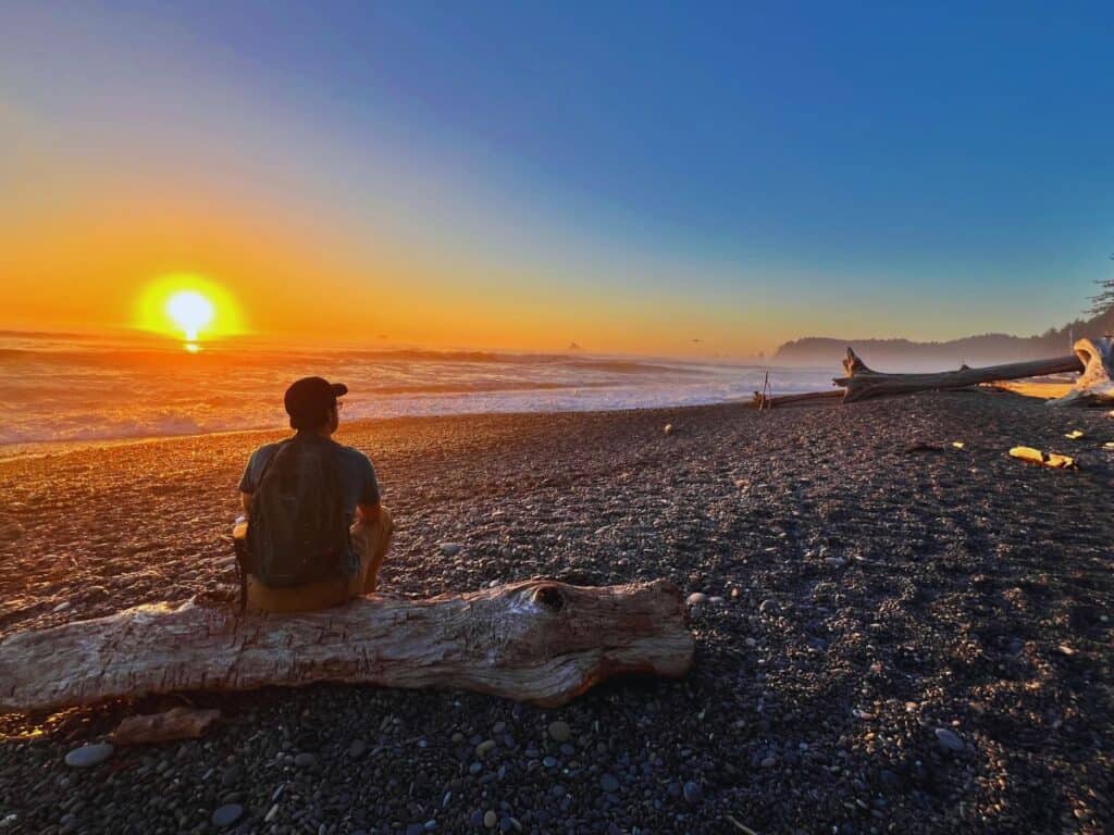 A man sits on a piece of driftwood to watch sunset at Rialto Beach, Olympic National Park, WA