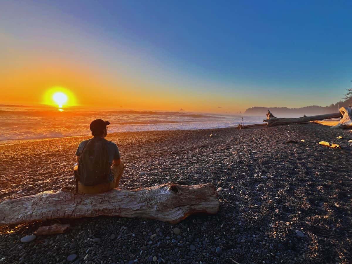 A man sits on a piece of driftwood to watch sunset at Rialto Beach, Olympic National Park, WA