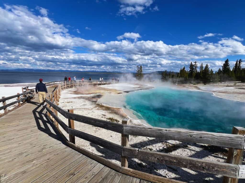 A man walks a boardwalk trail past a steaming blue pool in West Thumb Geyser Basin, Yellowstone National Park.