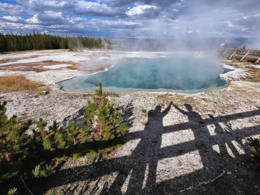 A man and woman perform a shadow high five in front of a blue spring in Yellowstone National Park, Wyoming