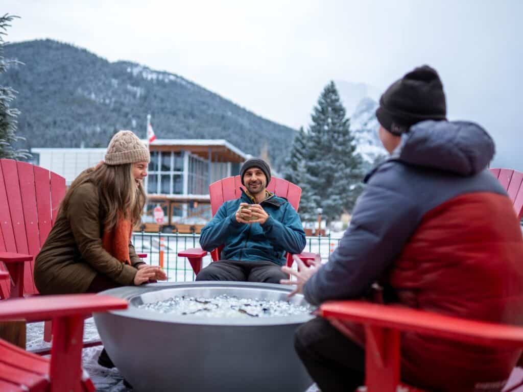 Three friends sit around a fire pit, sipping hot chocolate.
