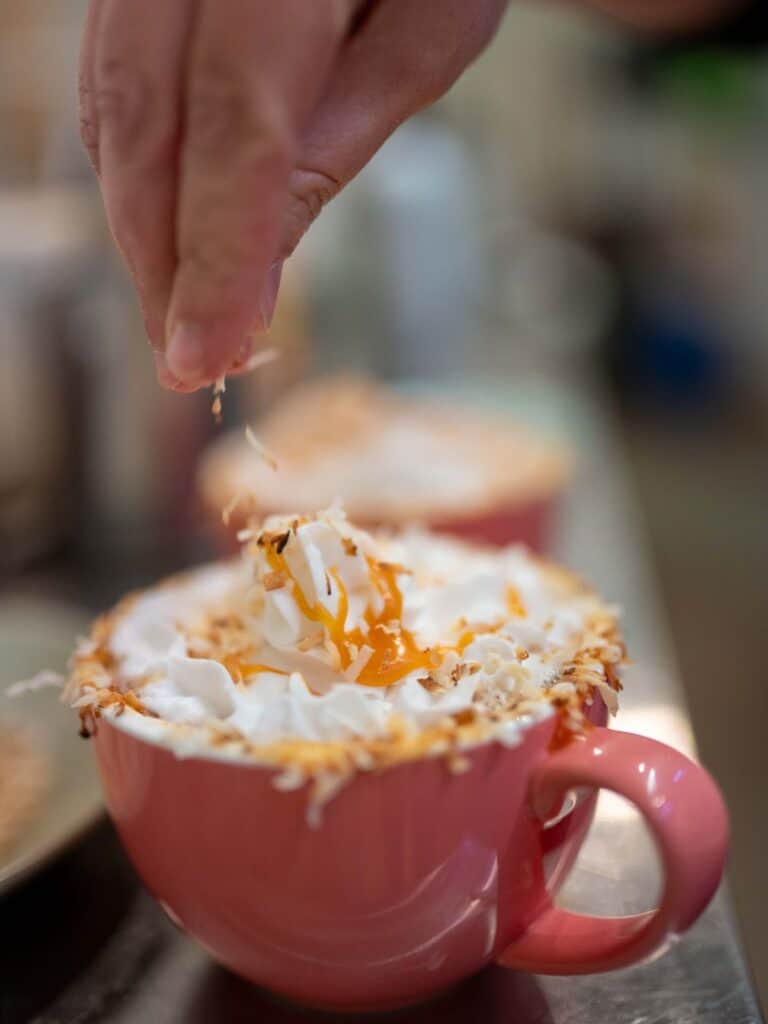 A hand sprinkles toppings on a mug of hot chocolate.