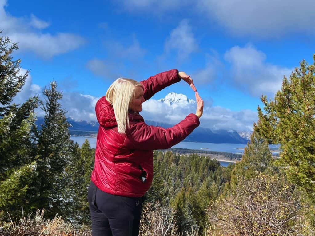 A woman hold her arms in a circle to highlight a snowy mountain peak on the horizon in Grand Teton National Park.
