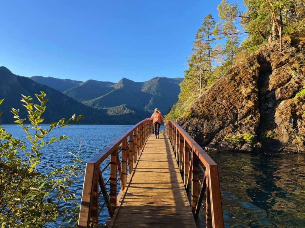 A woman walks across a footbridge with mountains in the background in Olympic National Park, WA.
