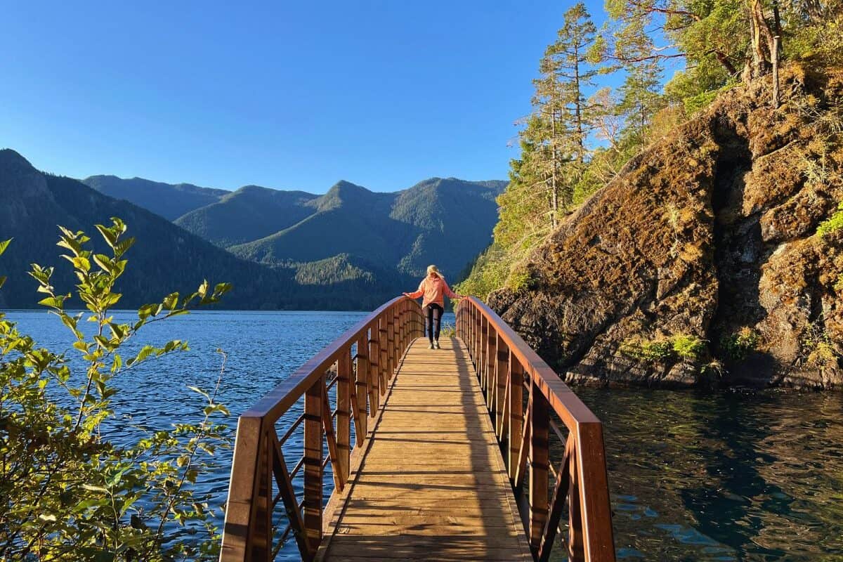 A woman walks across a footbridge with mountains in the background in Olympic National Park, WA.