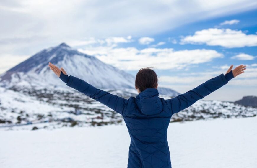 A woman faces a snowy mountain, holding up both arms in joy at the view.