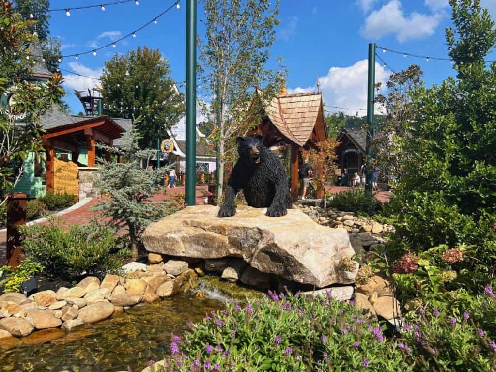 A black bear statue appears to climb a rock above a small stream surrounded by flowers and greenery.
