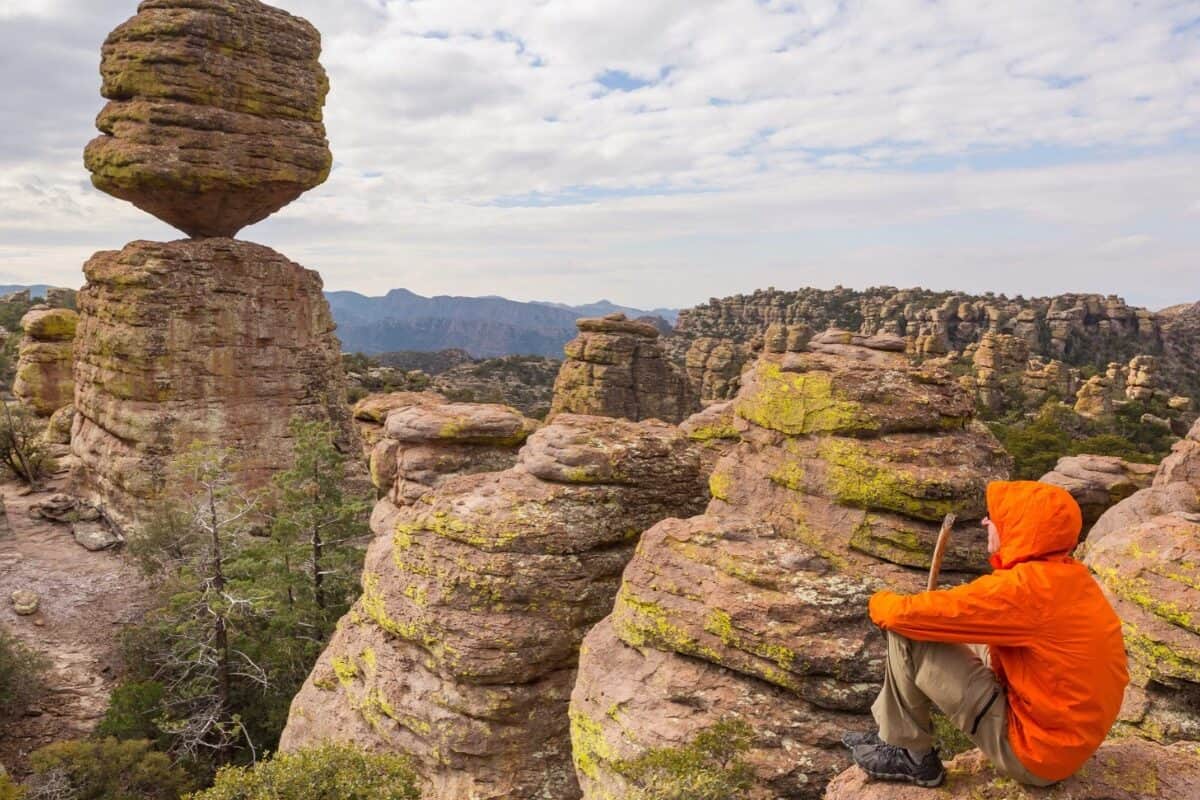 A hiker sits on a ledge overlooking a large rock balancing on a rocky column.