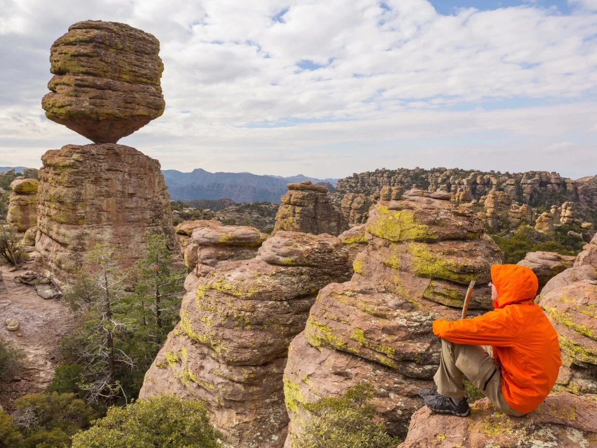 A hiker sits on a ledge overlooking a large rock balancing on a rocky column.